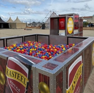 Ballenbak gevuld met rode, gele en blauwe ballen, omgeven door metalen en glazen panelen met reclame voor "Kasteel Rouge" bier, op een zandoppervlak tijdens een festival of evenement