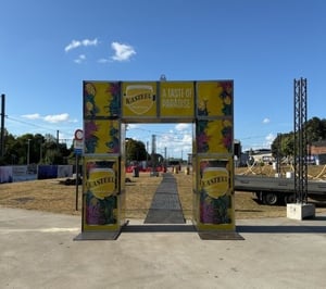 Buitenevenement met een grote gele boog met reclame voor "Kasteel Tropical" bier, voorzien van de tekst "A TASTE OF PARADISE" en afbeeldingen van bierblikjes, met een podium, tenten en apparatuur op de achtergrond onder een heldere lucht.