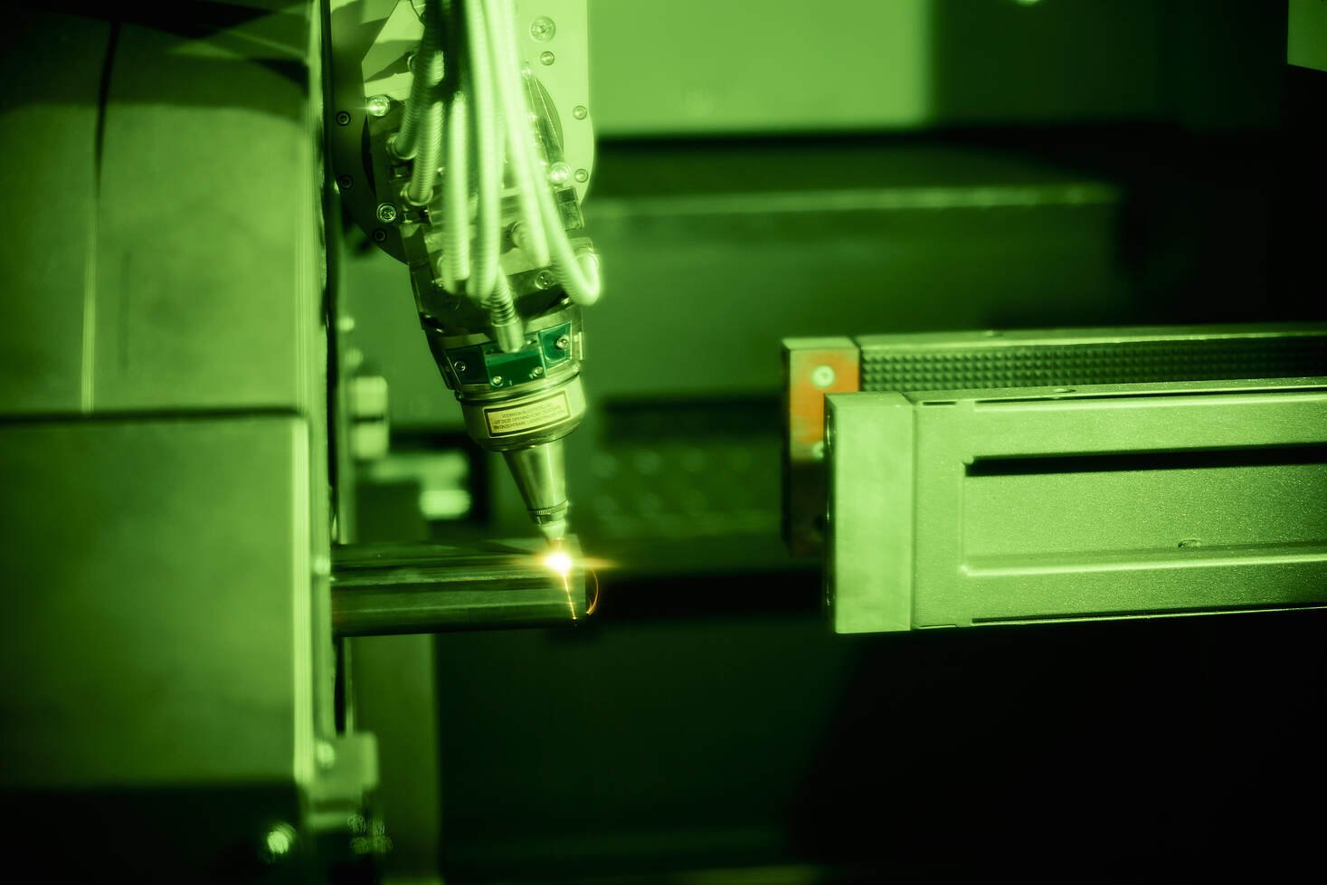 Close-up van een Adige LT-8.20 3D buizenlaser in werking, waarbij een geconcentreerde laserstraal metaal snijdt en vonken genereert. De industriële omgeving bevat diverse mechanische onderdelen en is groenachtig verlicht.