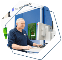 Paul bedient een TRUMPF lasersnijmachine in een moderne productieomgeving. Hij staat geconcentreerd achter een computerscherm, gekleed in een donkerblauwe werktrui met bedrijfslogo. Op de achtergrond is de TruLaser Center zichtbaar met groene veiligheidsramen, wat duidt op geavanceerde technologie en precisiewerk.
