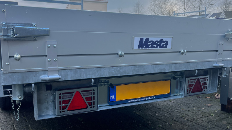 Achteraanzicht van een grijze metalen trailer van het merk Masta Aanhangwagens, Nederlandse kentekenplaat, rode reflectoren en metalen onderdelen, op een met stenen bestrate ondergrond met bomen en gebouwen op de achtergrond