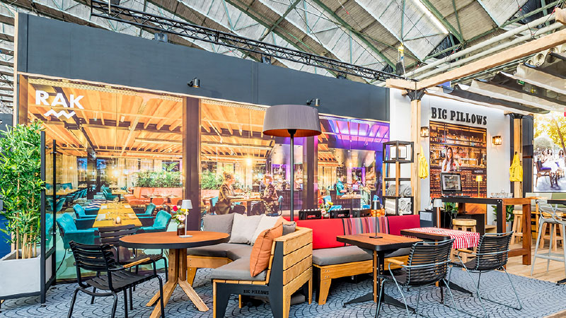 Gezellige en kleurrijke indoor zithoek van een restaurant of café met tafels, stoelen, een bank met kussens, grote vloerlamp en zicht op een bar met de naam 