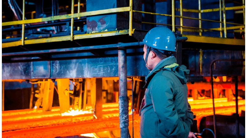 Persoon met blauwe helm en groene jas in een industriële omgeving met gloeiend hete staalbalken, typerend voor warmwalsprocessen.