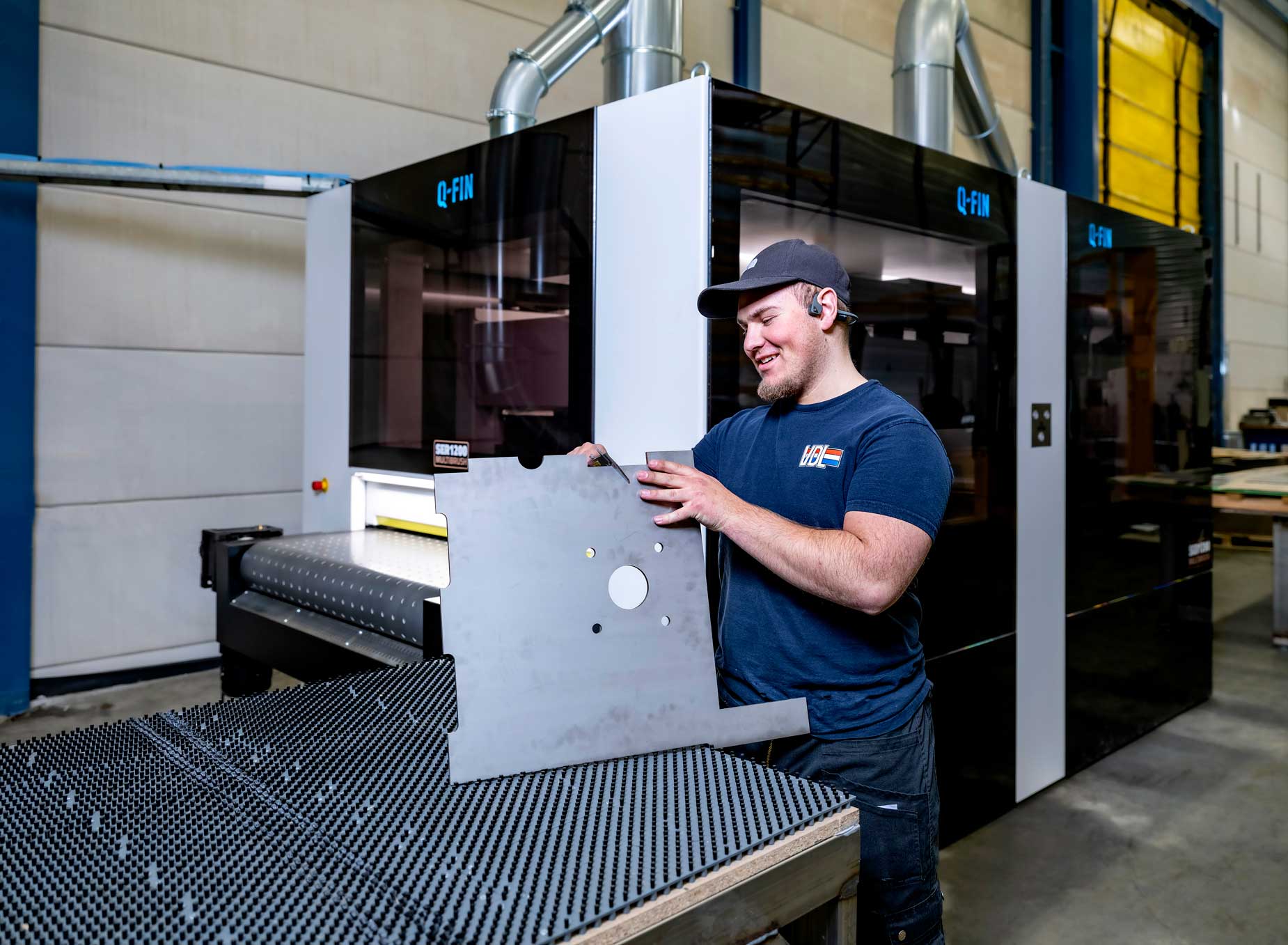 VDL-medewerker verwerkt een metalen plaat met uitsparingen naast een Q-FIN-machine, in een moderne productieomgeving ondersteund door OrderOn’s slimme orderverwerking.